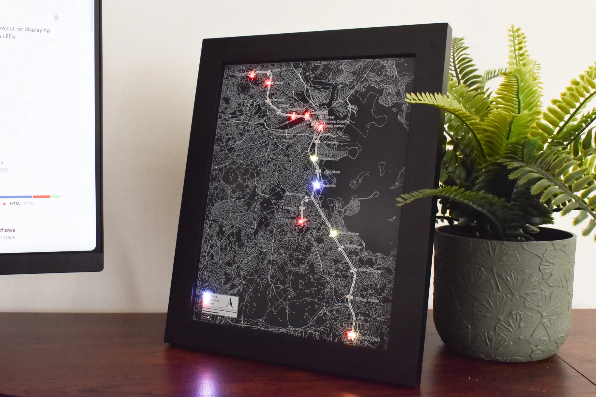 A framed CharlieBoard on a desk, displaying the MBTA Red Line with illuminated LEDs at several stations.
